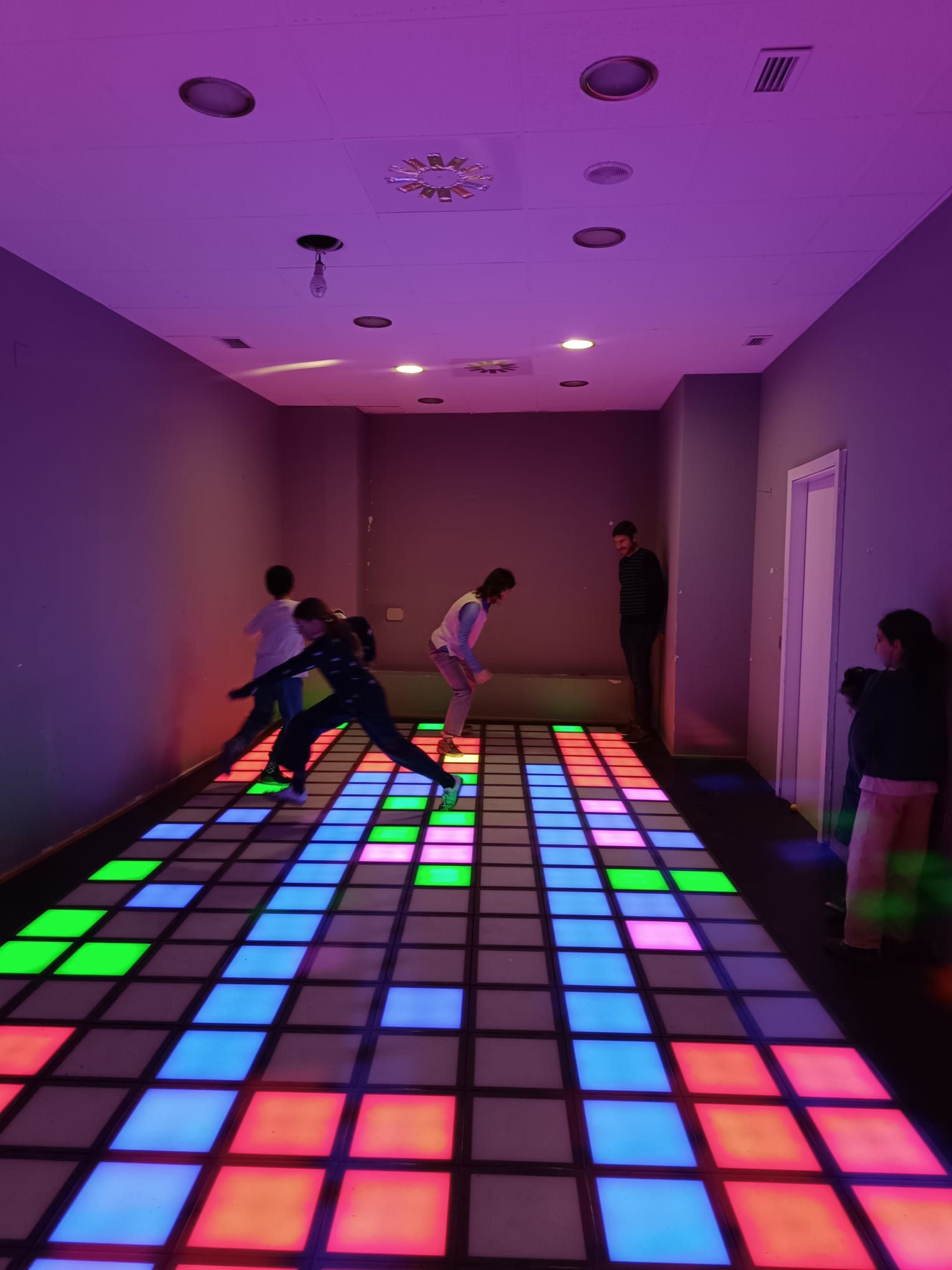Personas bailando sobre una pista multicolor iluminada en una sala tenue con iluminación púrpura
