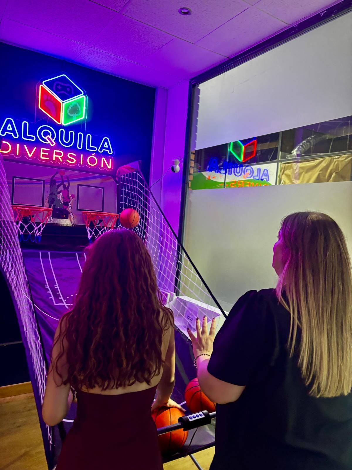 Dos personas están sentadas en la entrada de un salón recreativo cubierto, iluminada con luces de neón y con un letrero luminoso que dice «ALQUILA DIVERSION».
