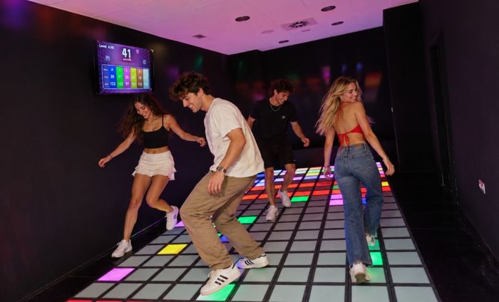 Personas bailando sobre un suelo de baldosas luminosas en una habitación tenue iluminada con luz rosa.