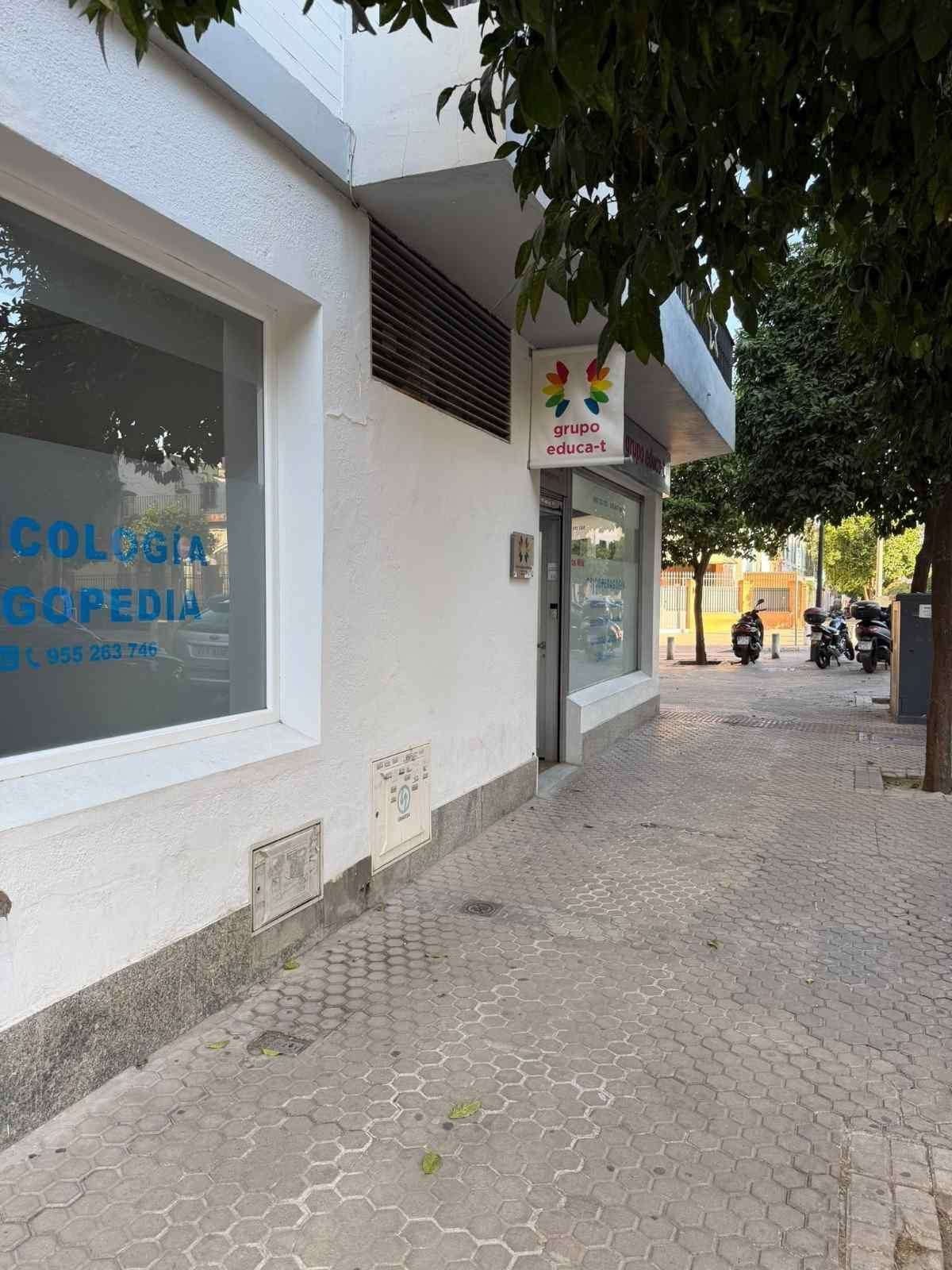 Acera frente a una clínica, con letreros azules, paredes blancas, árboles y algunas motocicletas estacionadas más adelante