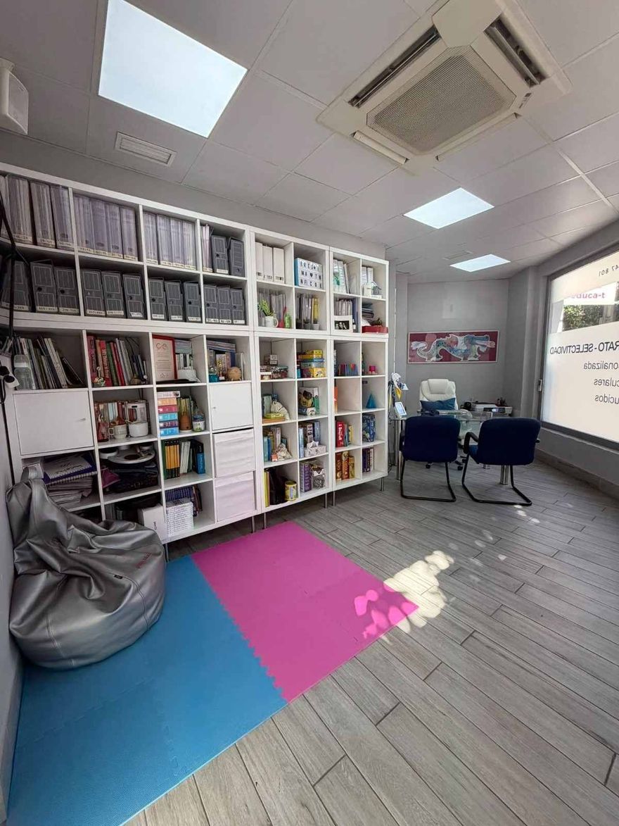 Sala de lectura luminosa con estanterías blancas, un puf, una alfombra rosa y azul y una mesita con sillas