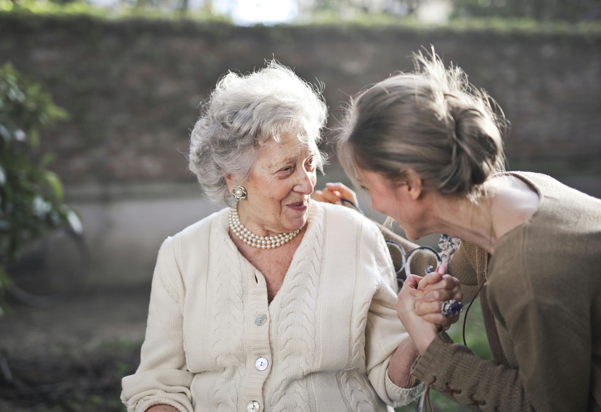 Una mujer ayuda a una anciana, ambas sonriendo afuera. La anciana lleva un cárdigan blanco y un collar de perlas.