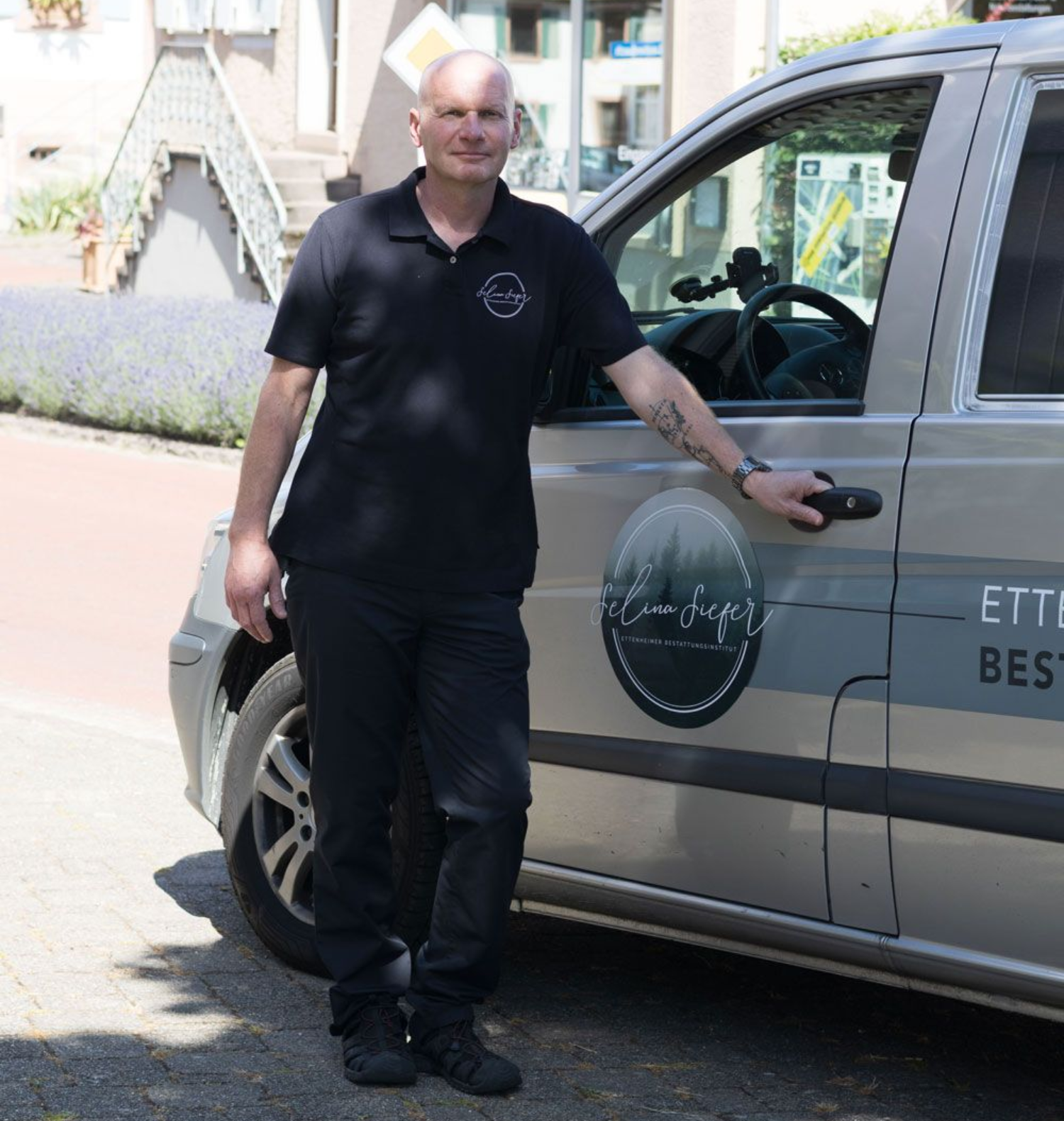 Ein Mann steht neben einem grauen Lieferwagen auf einer sonnigen Straße, die Hand am Türgriff.