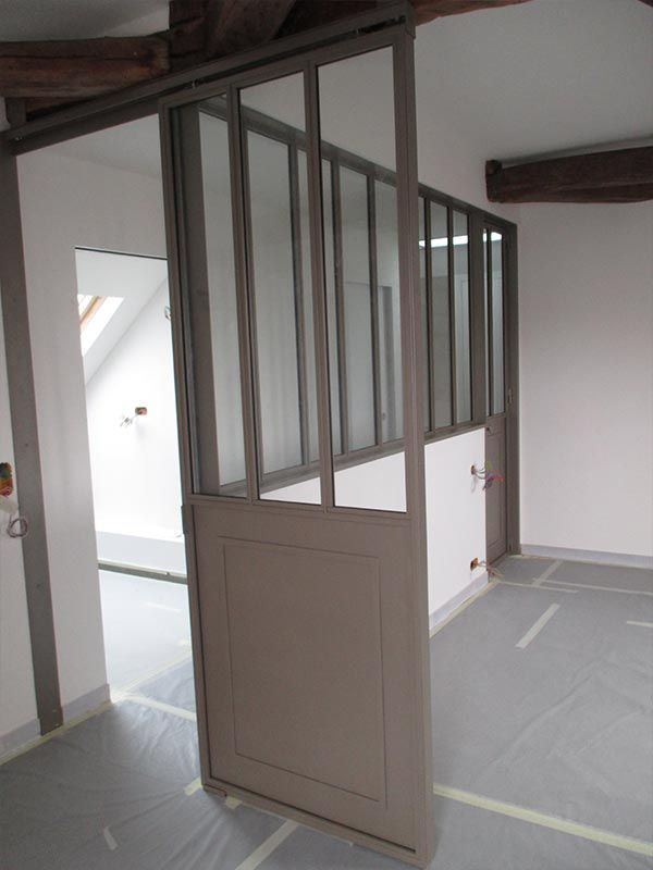 Une cloison de séparation avec une base en bois peint en gris et des panneaux supérieurs en verre à cadre métallique, installée dans une pièce non finie.