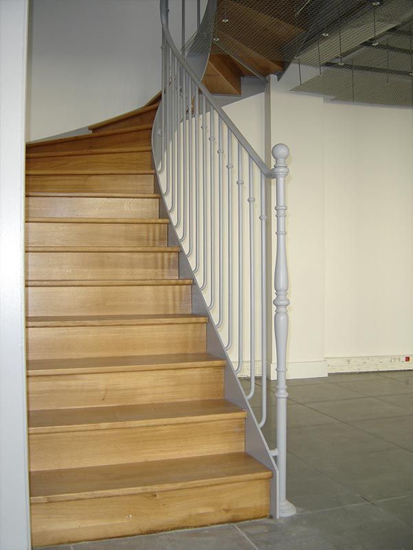 Un escalier en bois incurvé avec des marches claires et une rampe en métal gris dans une pièce au sol carrelé.