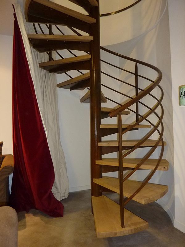 Dans une pièce, un escalier en colimaçon en bois avec une rampe en métal sombre se dresse à côté d'un rideau de velours rouge drapé.