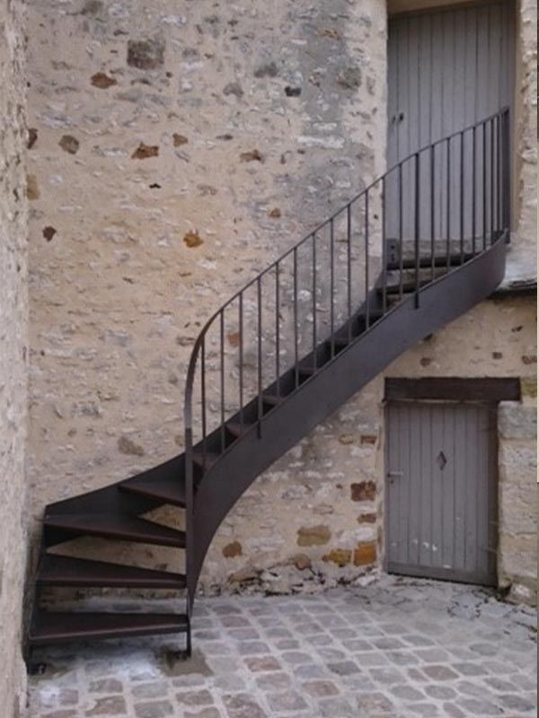 Un escalier étroit et incurvé en fer, doté de rampes verticales, mène à une porte en bois située à l'étage, contre un mur de pierre rustique.