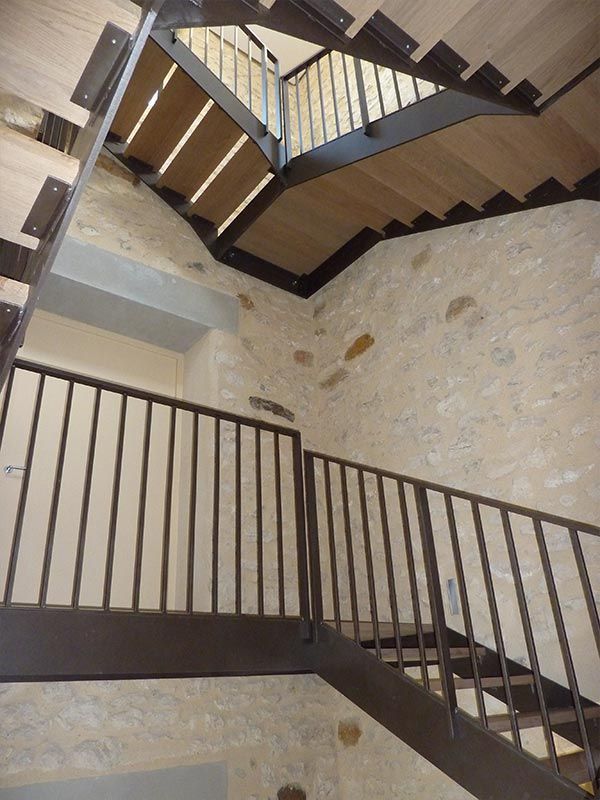 Vue intérieure d'un escalier moderne avec des rampes en métal foncé et des marches en bois clair, adossé à un mur en pierre texturée.