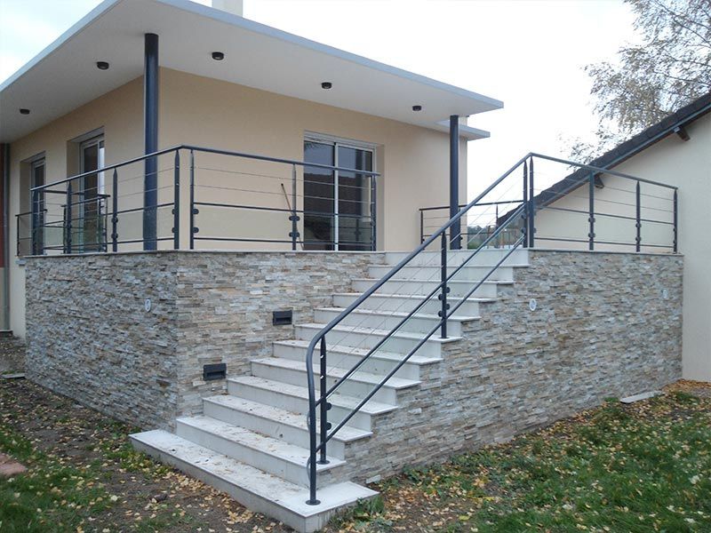 Une terrasse surélevée revêtue de pierre avec un escalier blanc, des balustrades en métal foncé et une façade de maison moderne.
