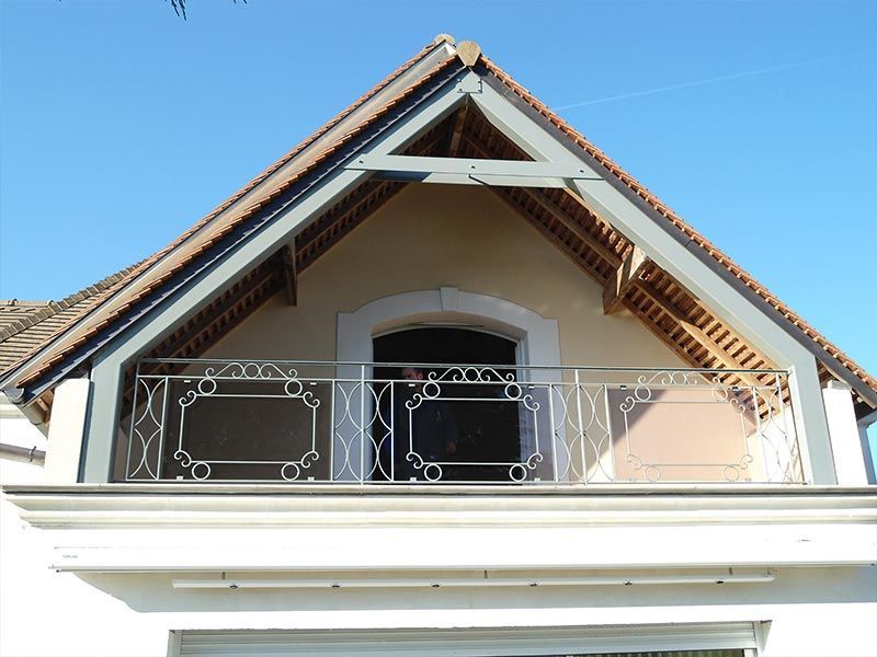 Un balcon avec une rambarde décorative en métal, situé sous le pignon d'une maison beige au toit de tuiles brunes.