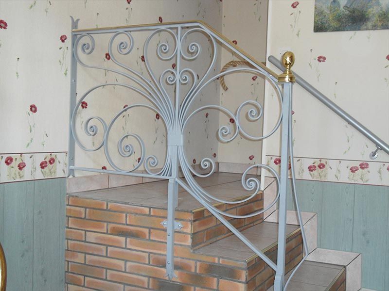 Rambarde décorative en fer forgé gris clair sur un escalier en briques, se détachant sur un papier peint à bordure florale.