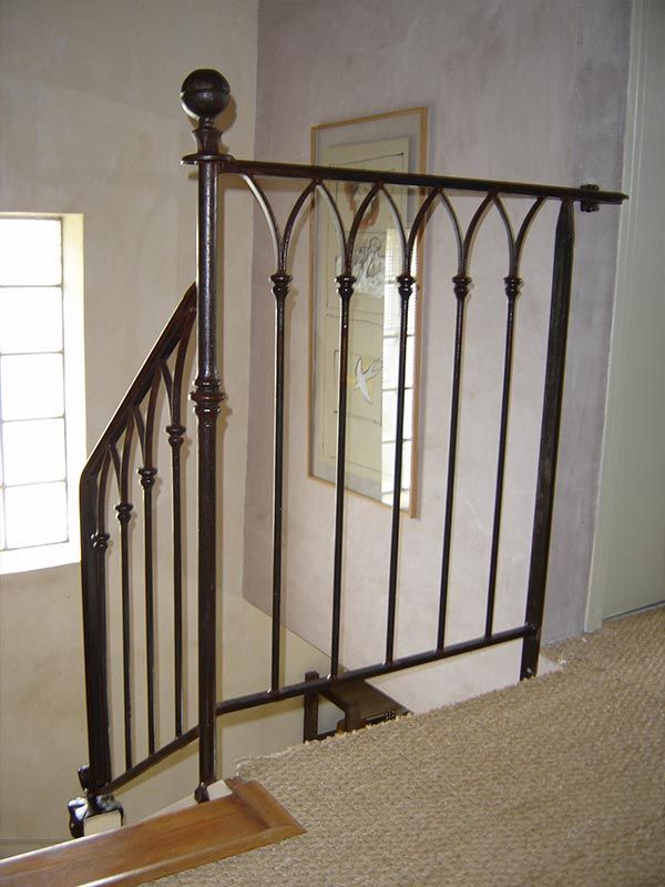 Une balustrade en fer forgé sombre, aux arcs gothiques, borde un escalier et un palier recouverts de moquette.