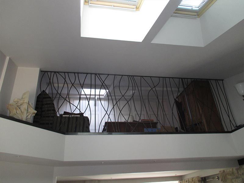 Une mezzanine est dotée d'une balustrade métallique moderne aux barres verticales ondulées, sous deux puits de lumière carrés.