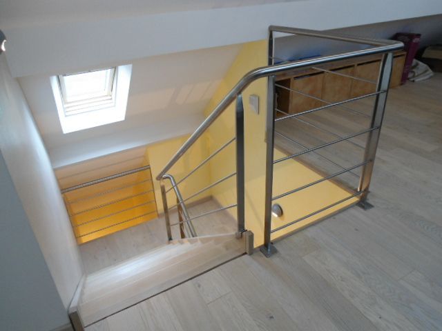 Escalier intérieur avec marches en bois, sol clair et rampe en acier inoxydable menant à un espace aux murs jaunes.
