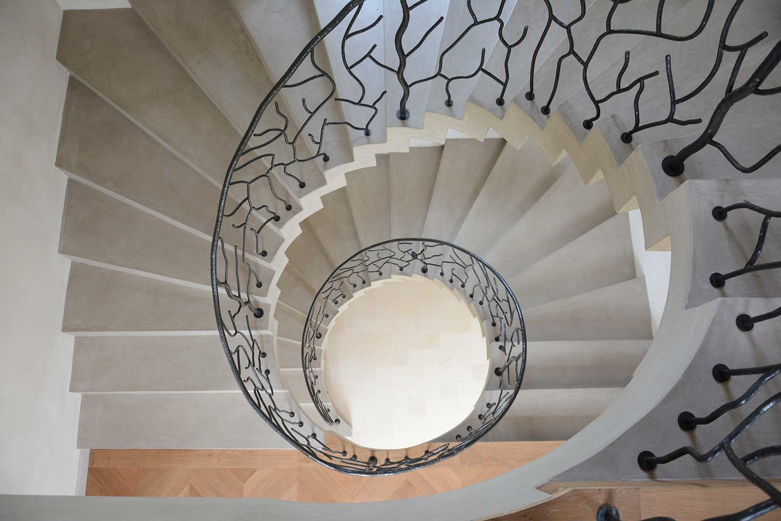 Vue en plongée d'un escalier en colimaçon aux marches en pierre claire et aux rampes en métal sombre ouvragées, évoquant des branches.