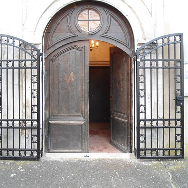 Deux portes doubles cintrées en bois sombre sont ouvertes, encadrées par des grilles en métal noir ouvragées, à l'entrée d'un bâtiment.