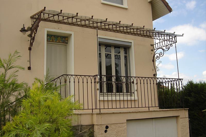 L'extérieur d'une maison beige présente un auvent en verre à armature métallique au-dessus d'une porte et une balustrade de balcon en fer forgé.