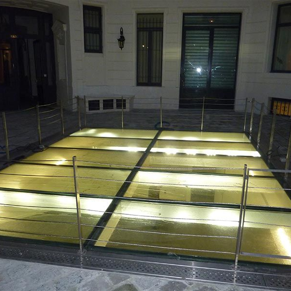 Une plateforme extérieure éclairée, au sol de verre et entourée de garde-corps métalliques, située devant un bâtiment la nuit.
