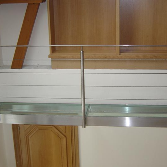 Une rambarde en acier inoxydable et une étagère en verre sont fixées contre un mur blanc, avec des boiseries et des meubles en arrière-plan.