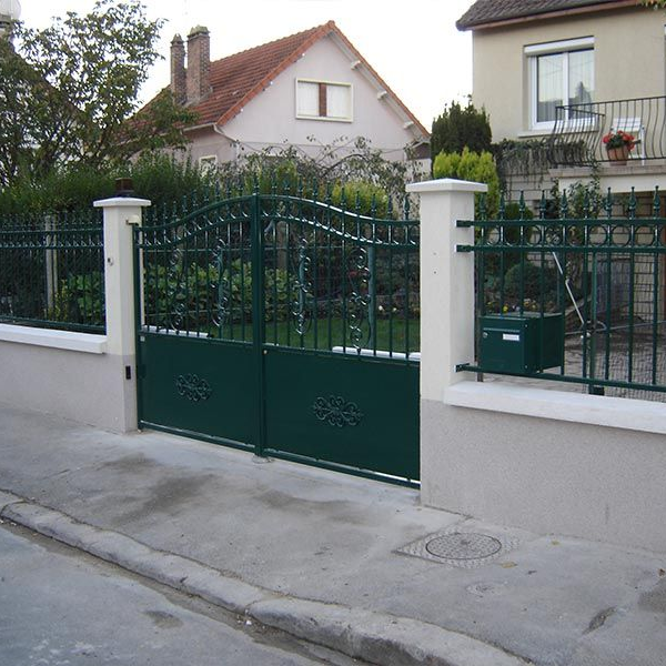Un portail en métal vert encastré dans un muret à piliers blancs, devant une maison d'habitation.