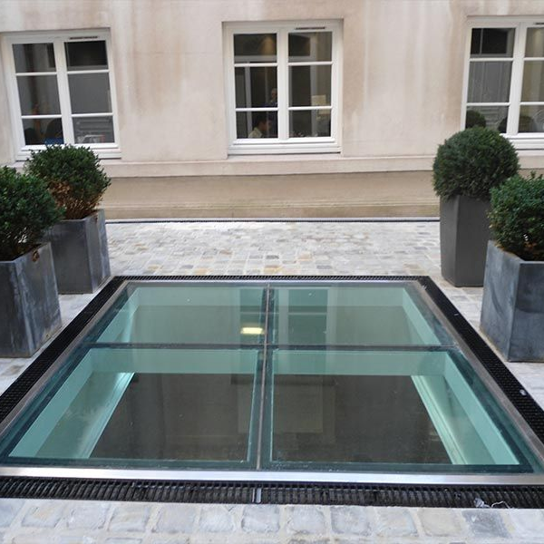 Un grand panneau de verre carré est encastré dans une cour pavée et entouré de quatre arbustes sphériques en pot.