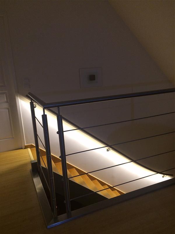 Un escalier avec des marches en bois et une rampe en métal, éclairé par un ruban LED à lumière chaude le long du mur.