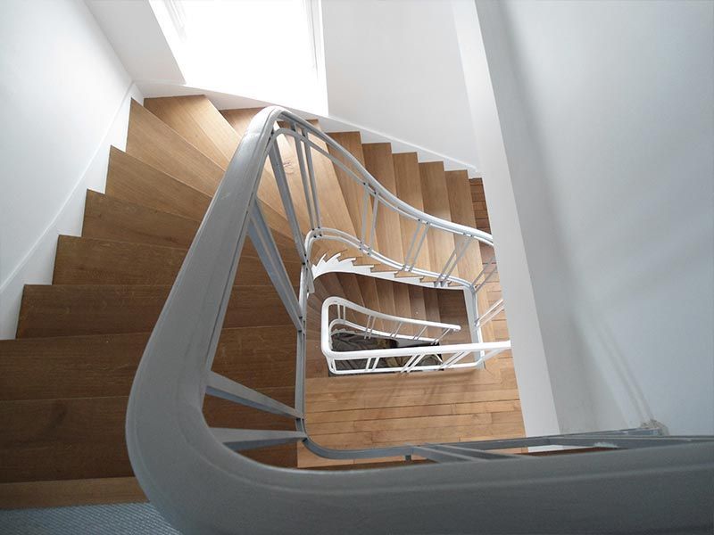 Vue en plongée d'un escalier moderne à rampe blanche et marches en bois, descendant en spirale vers une cage d'escalier lumineuse.