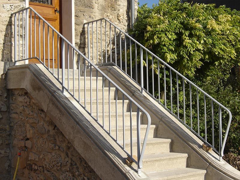 Un escalier en pierre mène à une porte en bois, avec des rampes en métal clair de chaque côté, à côté d'un mur en pierre et d'un buisson.