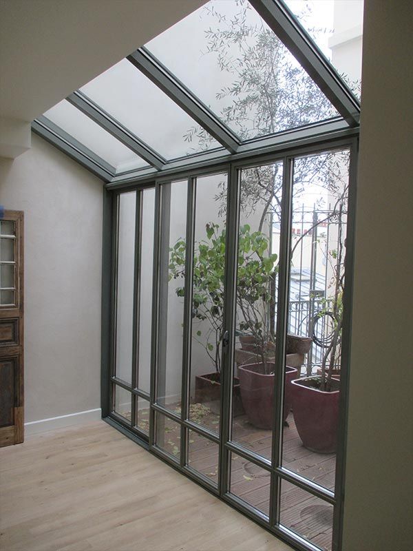 Une pièce avec des baies vitrées du sol au plafond et une verrière inclinée, donnant sur un petit patio avec des plantes en pot.