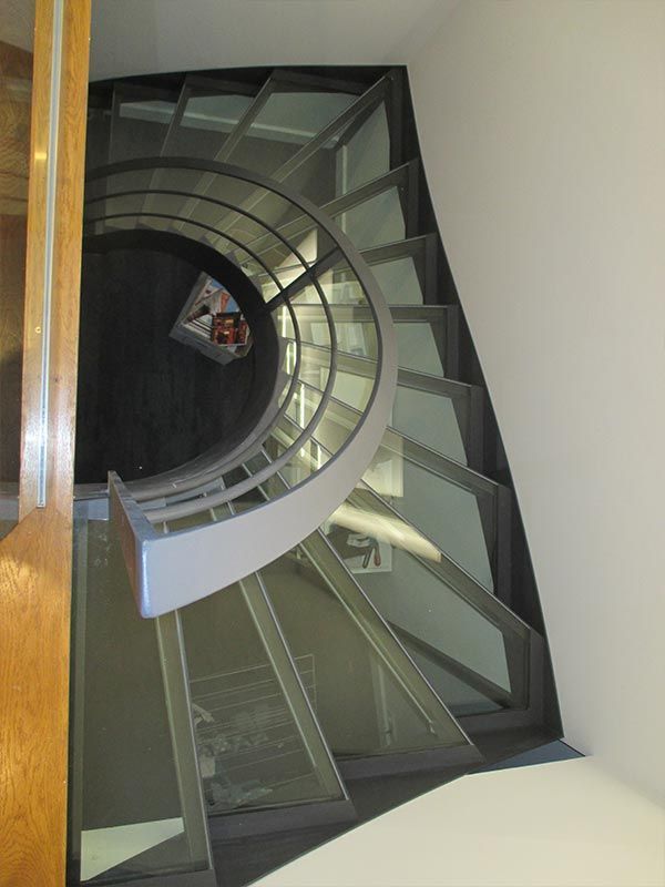 Un escalier en colimaçon avec des marches en verre et une rampe grise incurvée descendant d'un niveau supérieur.