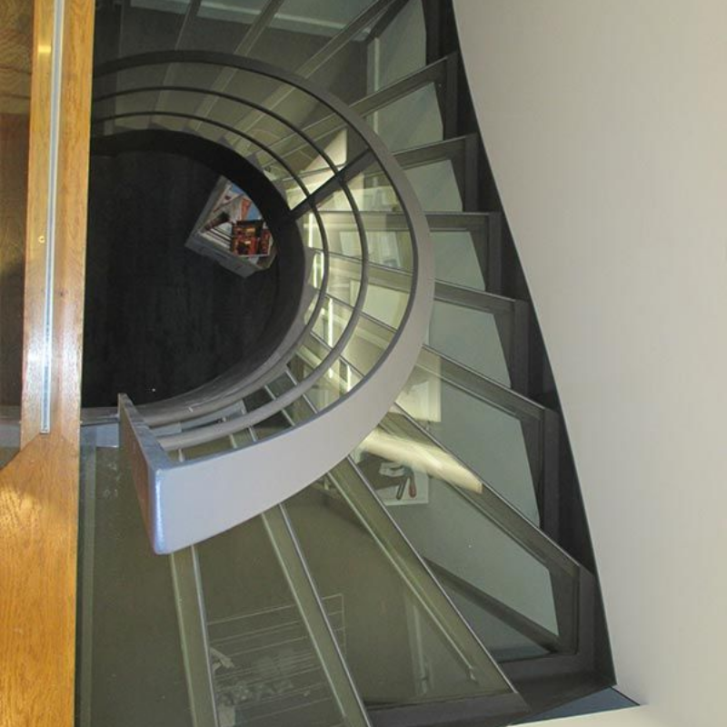 Vue plongeante d'un escalier en colimaçon avec des marches en verre et une rampe incurvée grise dans un intérieur moderne.
