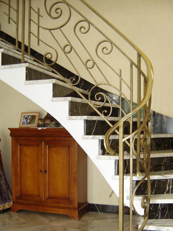 Dans un intérieur de maison, un escalier métallique incurvé, orné de rampes travaillées et de marches en marbre foncé, se trouve à côté d'une armoire en bois.