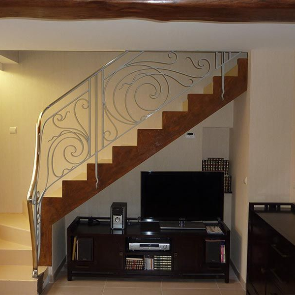 Un téléviseur et un meuble multimédia sont installés sous un escalier en bois avec une rampe en métal clair ornée.