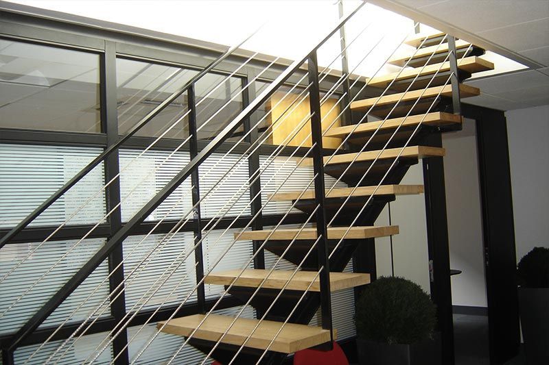 Escalier moderne à marches ouvertes avec marches en bois, structure en acier noir et garde-corps à câbles horizontaux dans un bureau professionnel.
