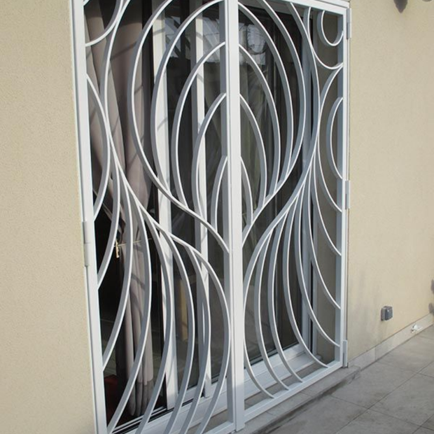 Grille de sécurité en métal blanc ornée d'un motif décoratif incurvé en forme de feuille, installée au-dessus d'une porte coulissante en verre.