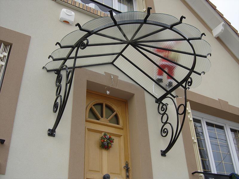 Un auvent décoratif en fer forgé noir, surmonté d'un toit en verre transparent incurvé, protège une porte en bois sur la façade beige d'une maison.