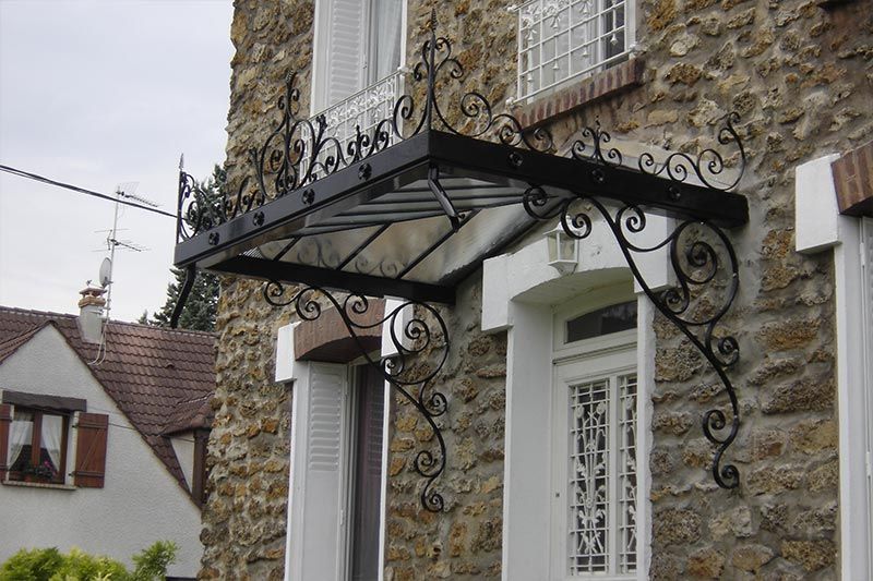 Un auvent de porte en fer forgé noir orné, avec des panneaux de verre, fixé à la façade rustique d'une maison en pierre.