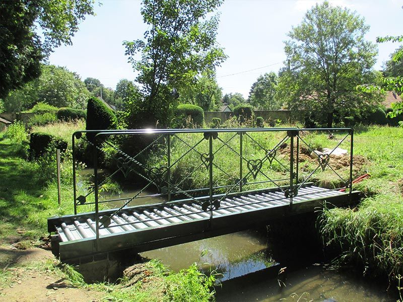 Une passerelle piétonne en métal vert enjambe un petit ruisseau paisible dans un parc verdoyant et ensoleillé.