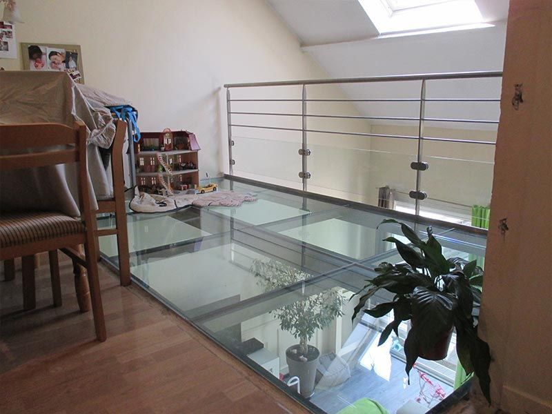 Une section de plancher en verre à l'étage supérieur, avec une rambarde en métal, surplombe une pièce inférieure avec une plante en pot.