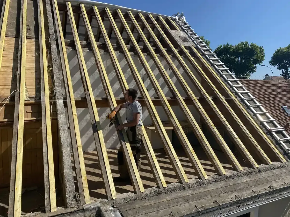 Un ouvrier installe des chevrons en bois sur un toit pentu d'une maison en construction.