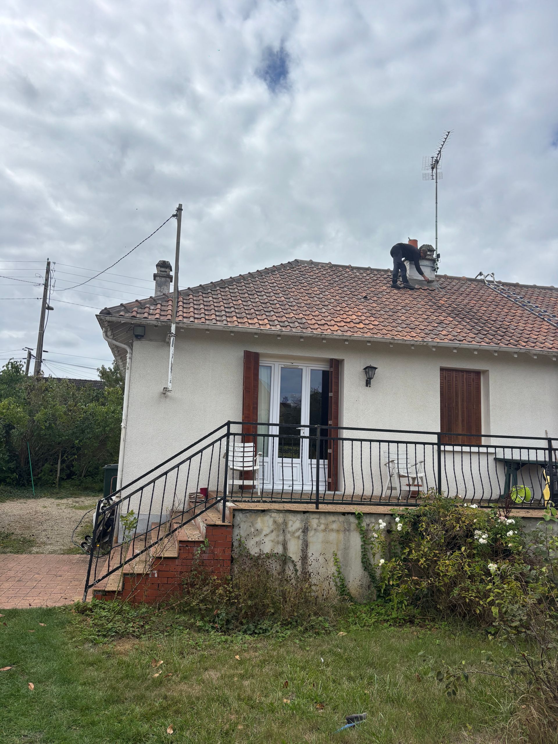 Petite maison blanche au toit de tuiles rouges, porche, escalier et ciel nuageux dans une cour rurale herbeuse.