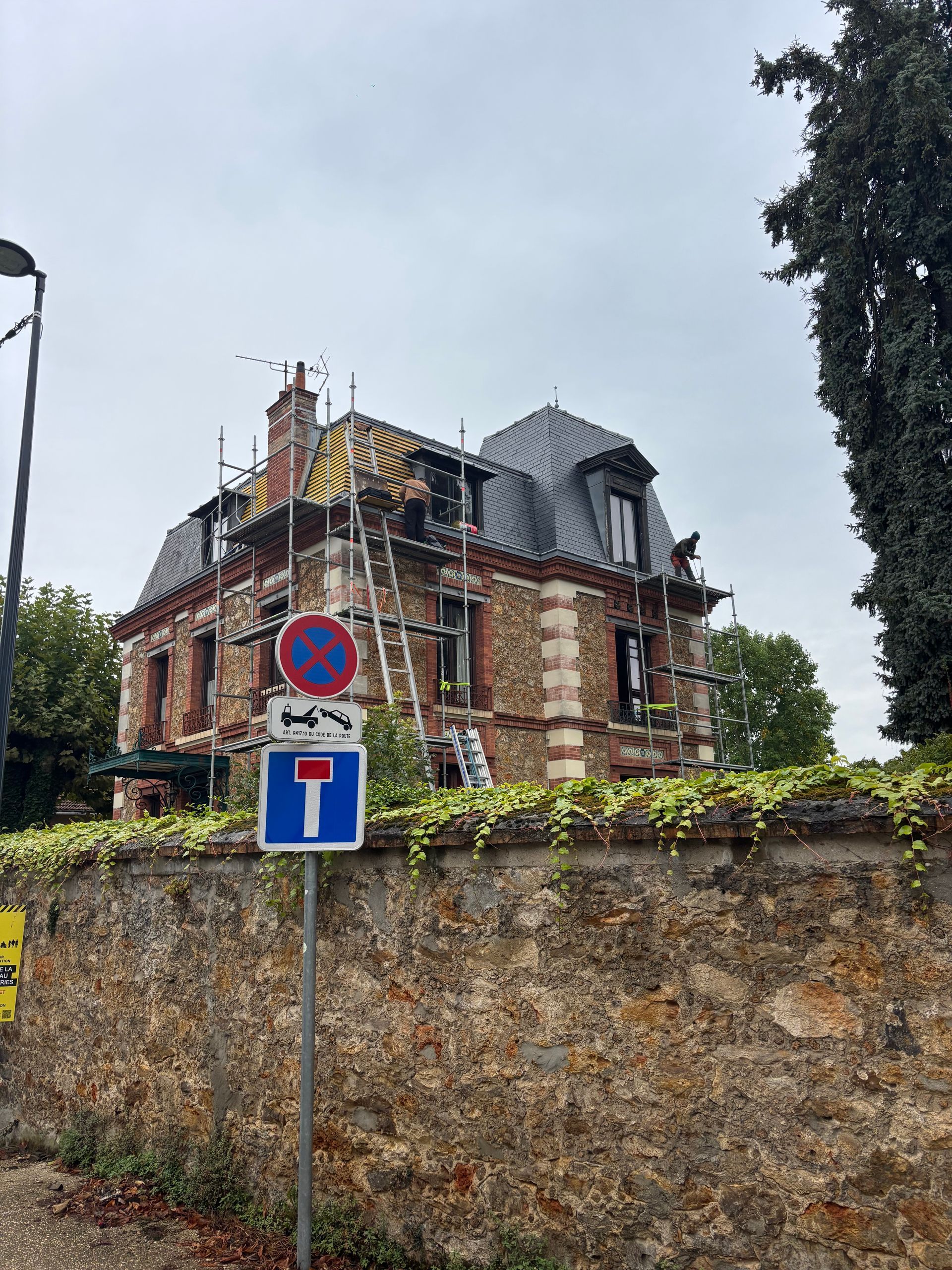 Vieille maison en briques en rénovation derrière un mur de pierre, sans parking et avec des panneaux indiquant une impasse.