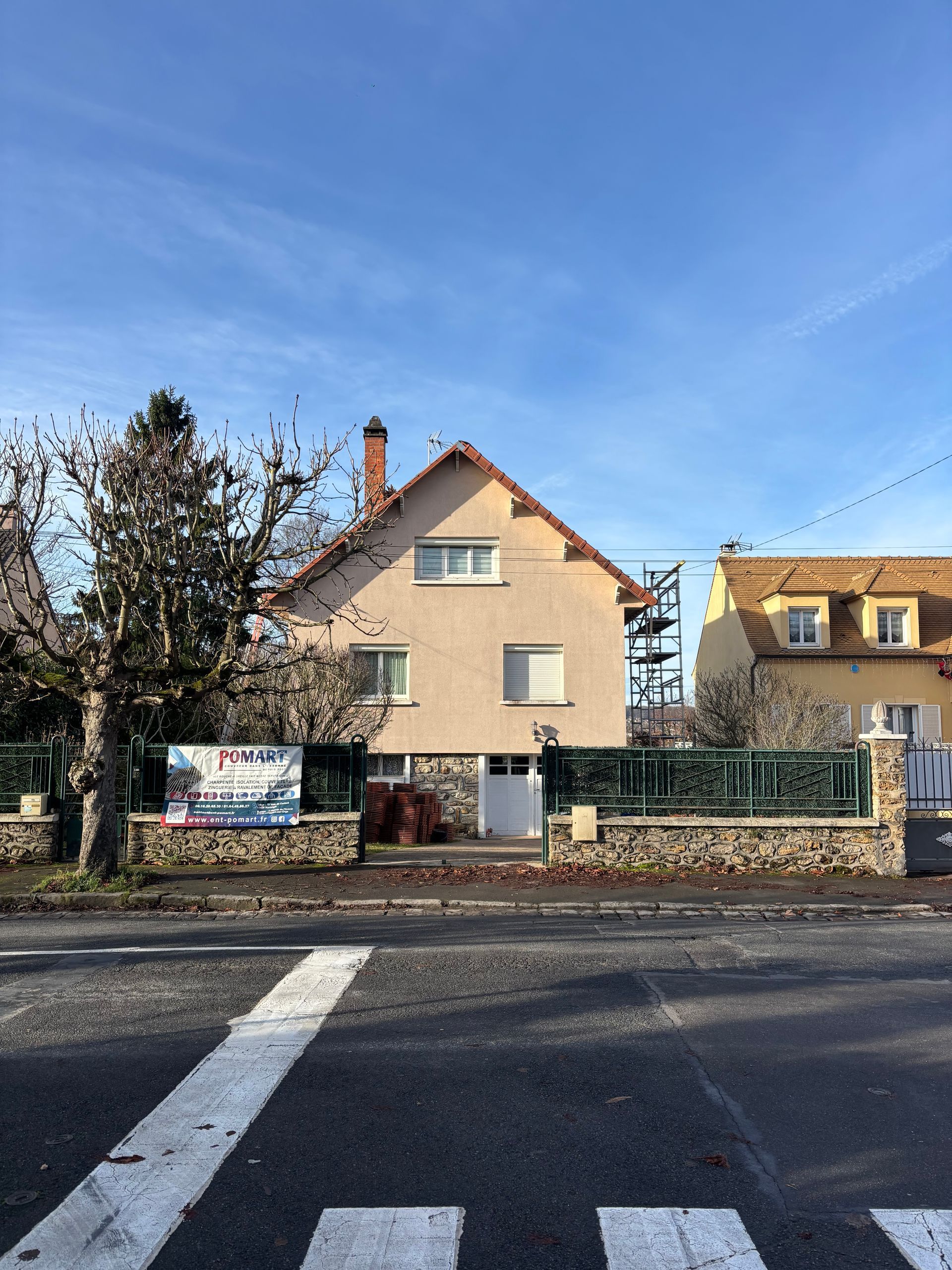 Rue résidentielle avec une maison, un mur de pierre, des arbres dénudés et un ciel bleu nuageux, vue depuis un passage piéton.