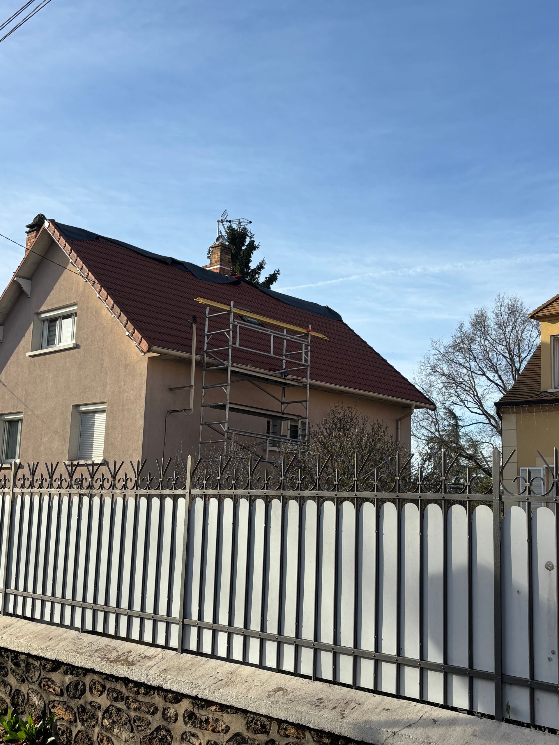 Maison beige au toit rouge derrière une clôture blanche, sous un ciel bleu azur.