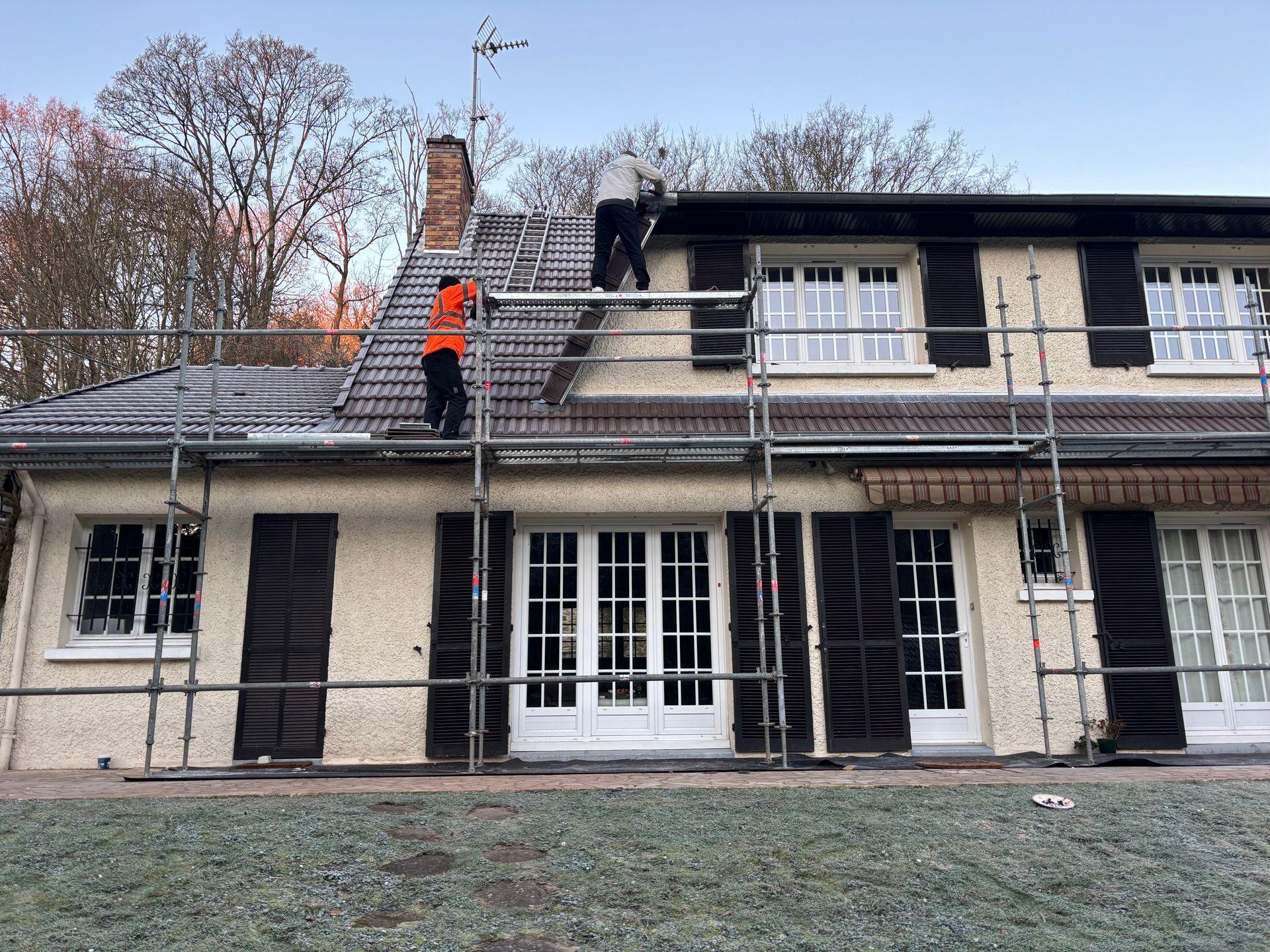 Deux ouvriers sur le toit d'un bâtiment beige aux fenêtres blanches et aux volets noirs installent un échafaudage.