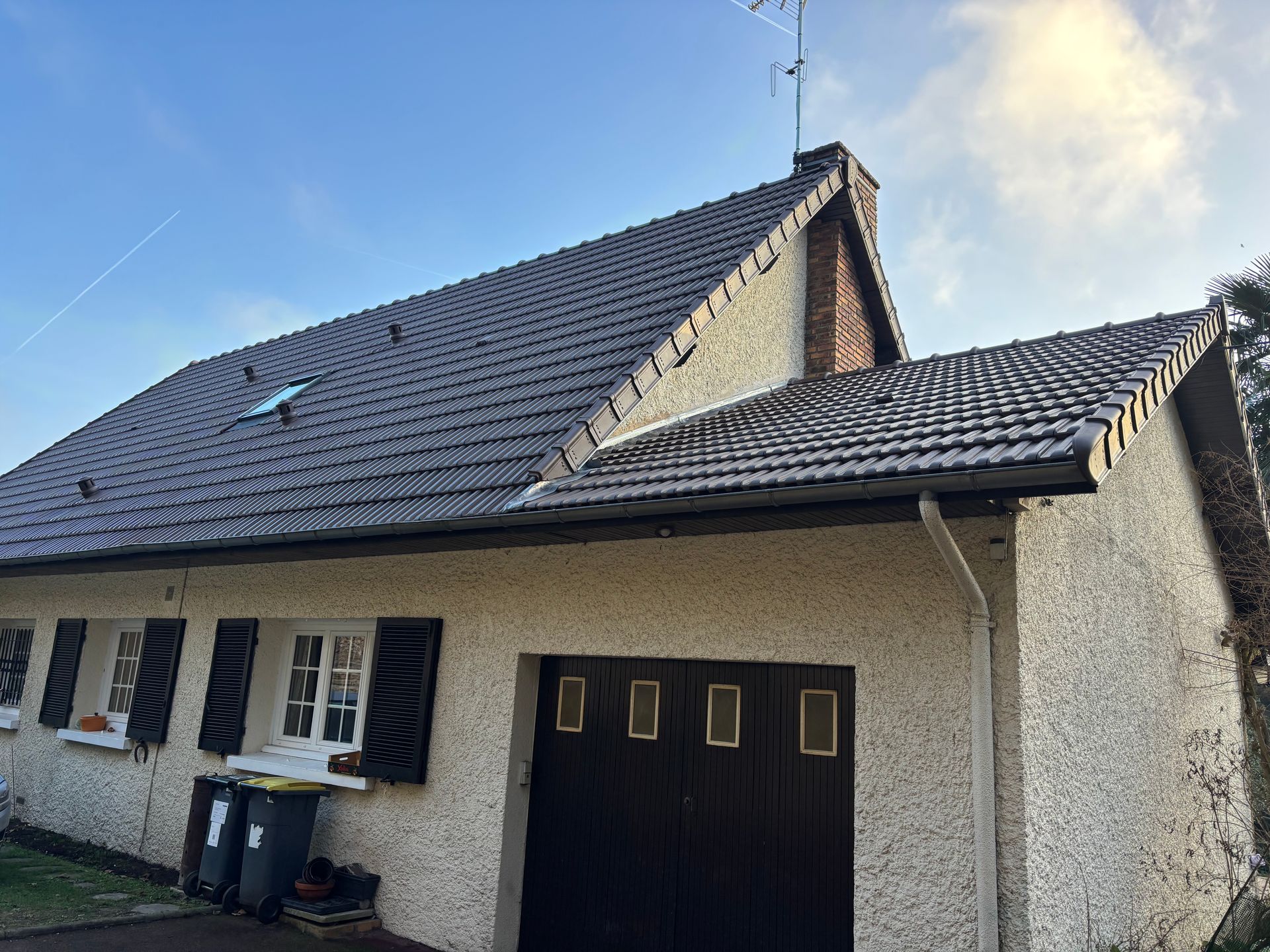 Petite maison beige au toit de tuiles foncées, fenêtres et porte de garage noire, sous un ciel bleu.