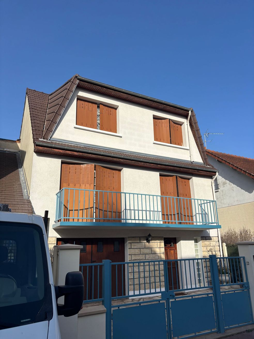 Maison à deux étages avec façade blanche, volets marron, balcon et portail bleus sous un ciel d'un bleu limpide.