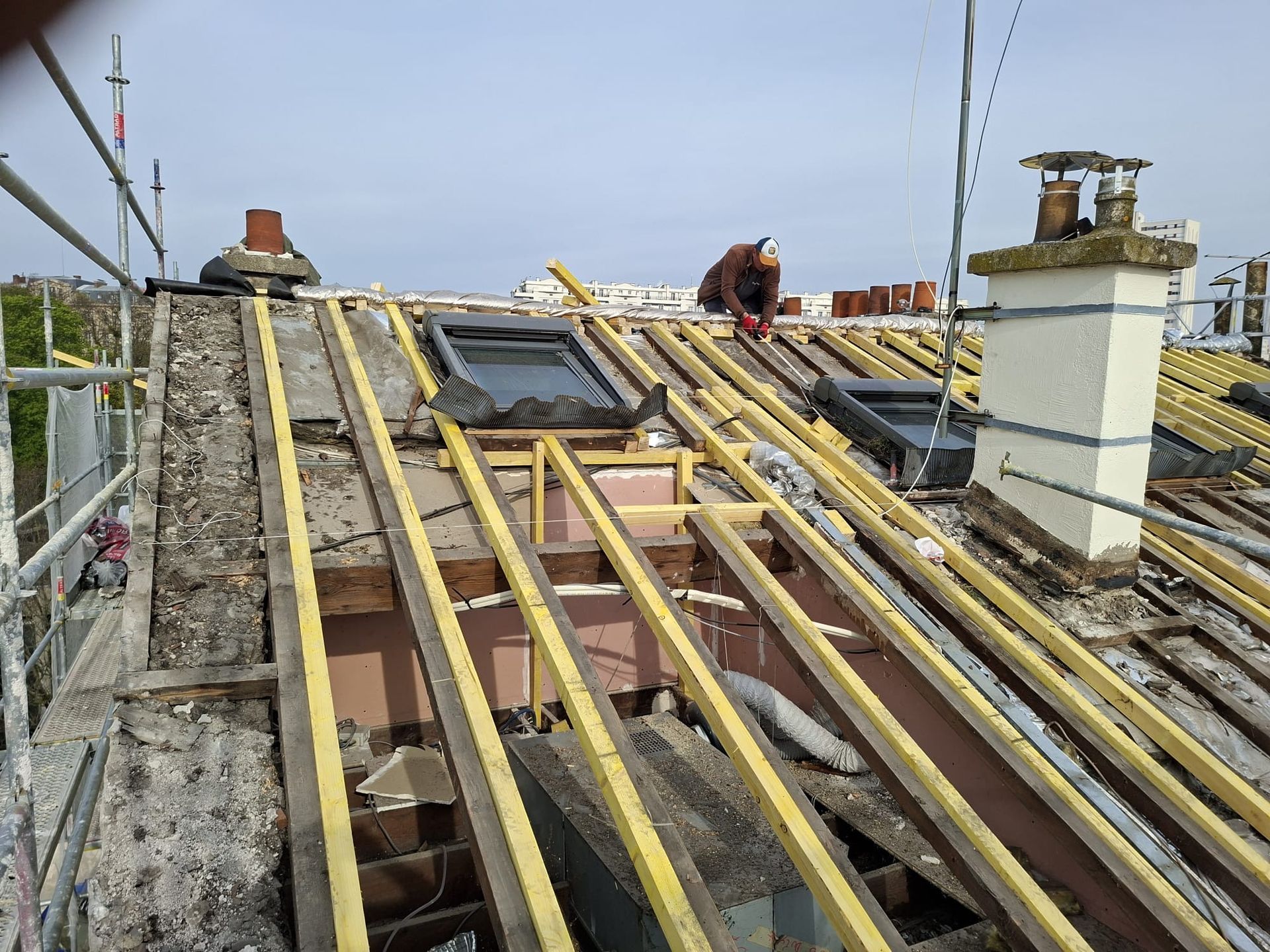 Toiture en construction avec poutres en bois, deux ouvriers et une lucarne.