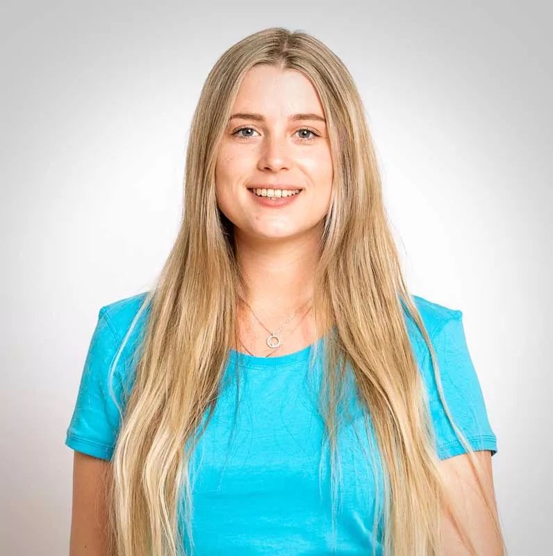 Eine Frau mit langen blonden Haaren trägt ein blaues T-Shirt und lächelt.