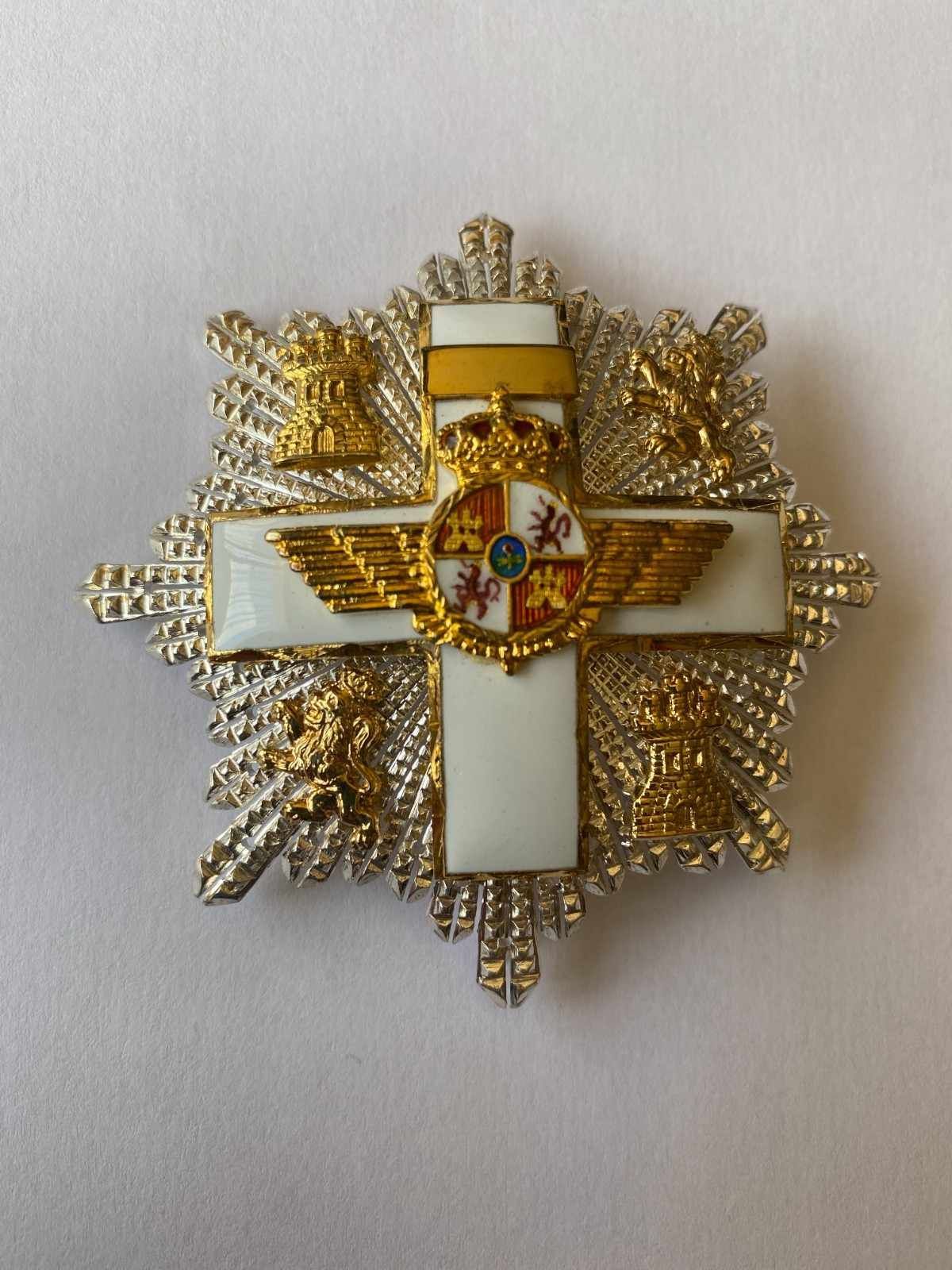 Medalla en forma de cruz decorativa de oro y blanco con un emblema central sobre fondo claro.